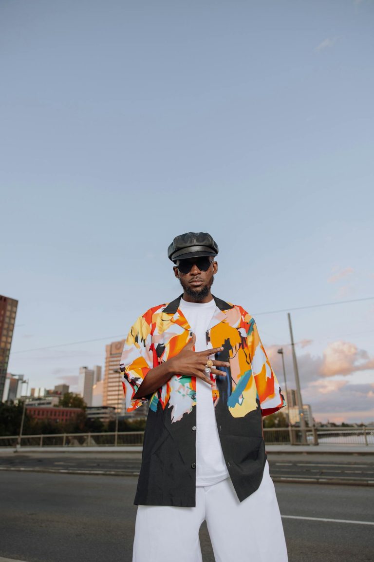 Stylish man in colorful shirt and hat poses on city street at sunset.