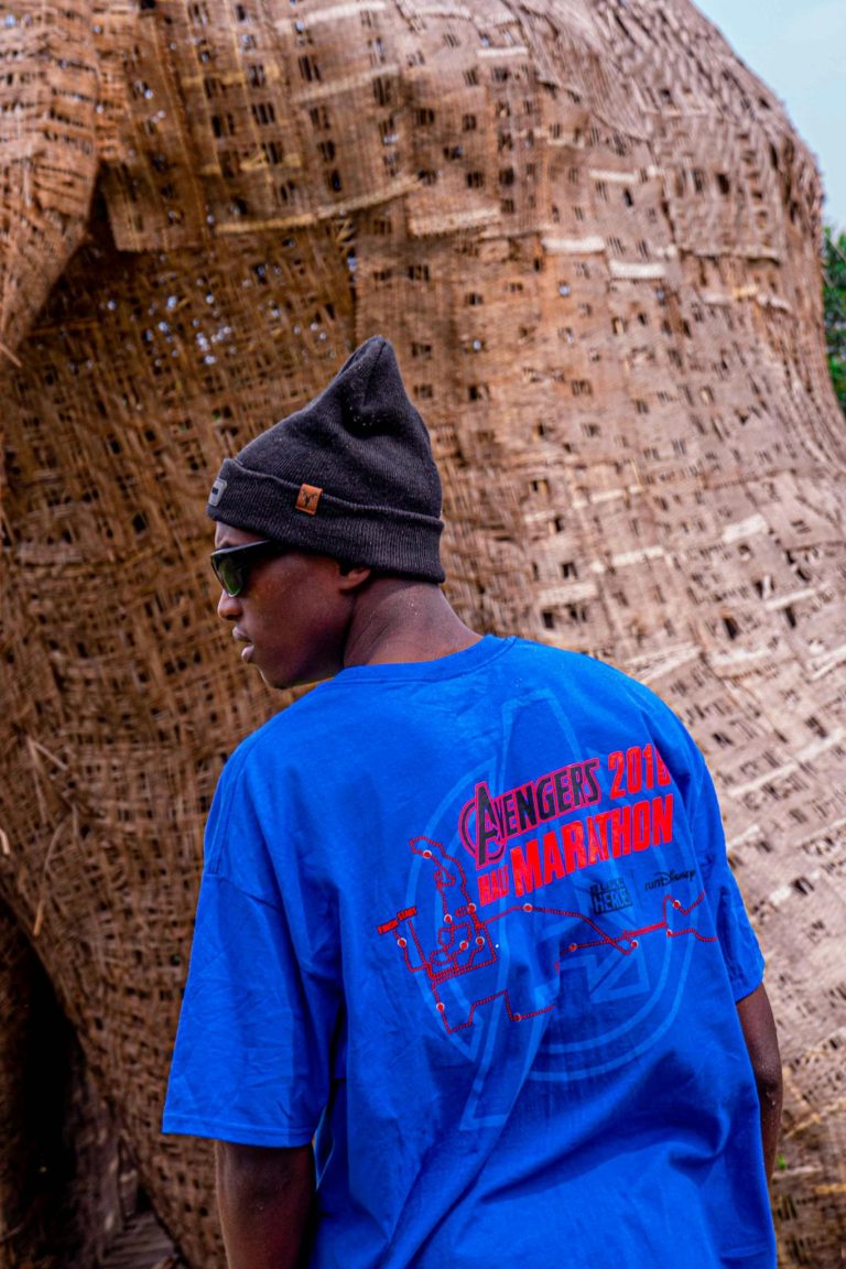 Stylish man wearing sunglasses and a blue t-shirt with Avengers logo standing outside.