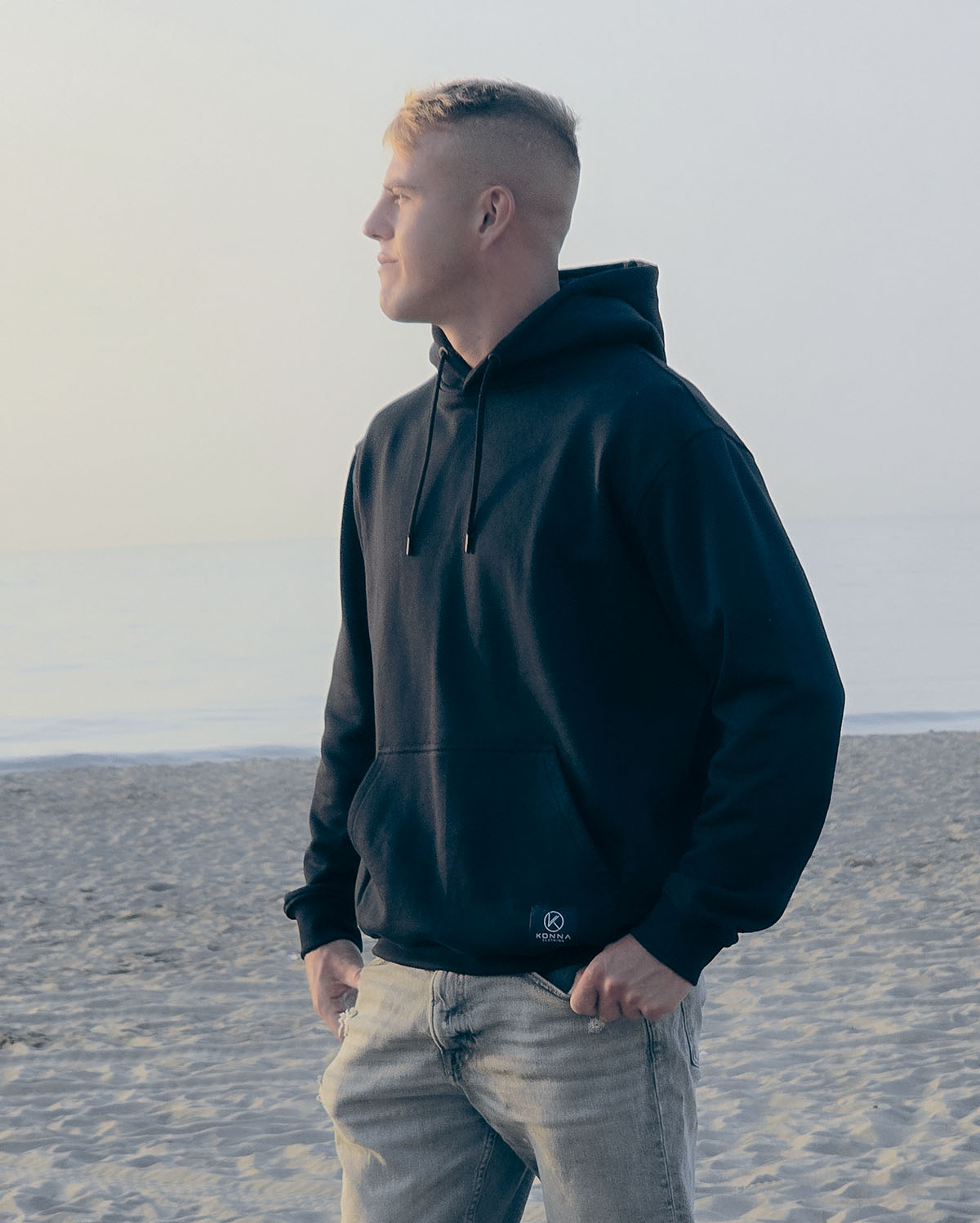 Young man wearing the black Konna Minimal Premium hoodie, standing on a sandy beach looking out towards the sea.